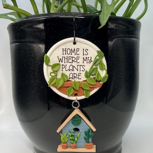 May include: A black ceramic plant pot with a decorative plant tag. The tag reads "HOME IS WHERE MY PLANTS ARE" and features a plant design. A small birdhouse charm with potted plants hangs below.