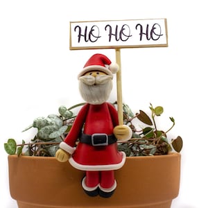 May include: A clay figurine of Santa Claus holding a sign that says "HO HO HO" sitting on the edge of a terracotta pot with a green plant.
