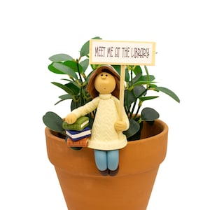 May include: A terracotta plant pot containing a green leafy plant and a figurine of a person with brown hair wearing a cream sweater and blue pants. The figurine holds a stack of three books and a sign that reads MEET ME AT THE LIBRARY.