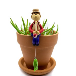 May include: A clay figurine of a person wearing a brown hat and red shirt, sitting on the edge of a terracotta pot with green plants. The person is holding a fishing rod with a red bobber and a green fish on the end of the line.