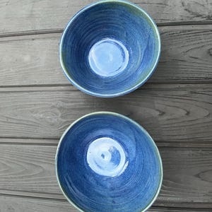 May include: Two round ceramic bowls with a glossy finish, featuring a deep blue colour with subtle variations and hints of green along the curved rims. The interior surfaces show visible brush strokes and a smooth, reflective texture throughout.