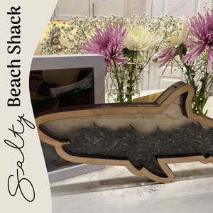 May include: A wooden shark-shaped decorative piece with a dark interior, possibly resin. The piece is on a white surface, with the text "Salty Beach Shack" on the left side. Flowers in vases are in the background.