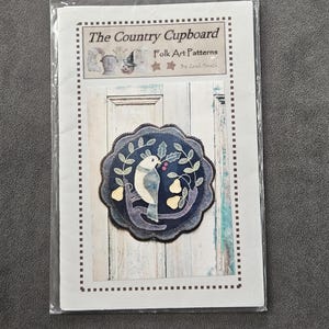 May include: A craft pattern packet titled "The Country Cupboard Folk Art Patterns" featuring a circular applique design. The applique depicts a bird perched on a branch with pears and leaves, in shades of blue, white, and yellow.