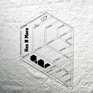 May include: A clear acrylic quilting ruler labeled Hex N More by Jaybird Quilts. The tool features black printed markings, including various geometric shapes like hexagons, half hexagons, jewels, and triangles with specific inch measurements.