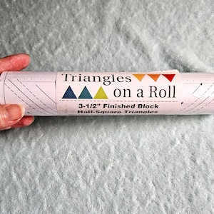 May include: A roll of light pink grid paper for quilting, labeled Triangles on a Roll, featuring a 3-1/2 inch finished block half-square triangle design. The paper has printed black dashed lines and arrows for sewing guidance.