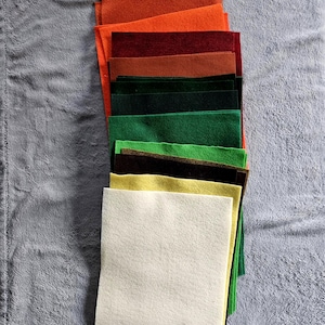 May include: A stack of colorful felt squares in shades of orange, burgundy, green, and cream. The felt squares are arranged in a fan shape, showcasing the variety of colors. The felt is soft and appears to be of high quality, suitable for crafting projects.