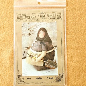 May include: A doll named Nellie, 7 inches tall, is featured in a clear plastic package. The doll is dressed in a brown and tan outfit, holding a small shovel and hoe. The package reads "Threads That Bind" and includes the number #121.