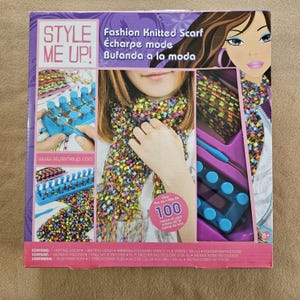 May include: A boxed craft kit labeled "STYLE ME UP!" for making a fashion knitted scarf. The box features images of the scarf, knitting tools, and the text "Fashion Knitted Scarf" in multiple languages. Includes over 100 meters of yarn.