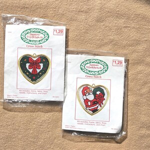 May include: Two Christmas cross-stitch kits in plastic packaging. Each kit features a heart-shaped design with a gold-colored frame. One kit shows a red bow on a green background, and the other depicts Santa Claus.