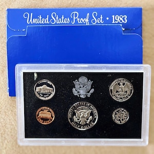May include: A 1983 United States Proof Set in a clear plastic case, with a blue cardboard sleeve. The set includes various coins, such as a penny, half dollar, and quarter dollar. The blue sleeve has the text "United States Proof Set • 1983".