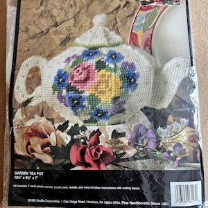 May include: A Garden Tea Pot kit. The kit includes a white teapot with a floral design in pink, blue, and yellow. The teapot measures 10.5" x 6.5" x 7". The kit includes plastic canvas, yarn, a needle, and instructions.