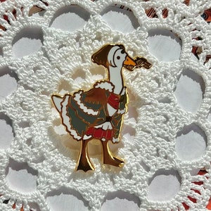 May include: A gold-coloured enamel pin of a goose wearing a green and brown coat and a red scarf. The goose is holding a small, gold-coloured trumpet in its beak.
