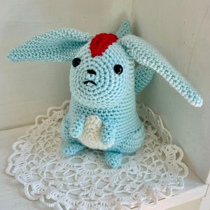Puede incluir: Un peluche de ganchillo hecho a mano con forma de criatura de orejas largas. El juguete es principalmente azul claro con una cresta roja y ojos negros. Se encuentra sobre un tapete blanco de encaje, mostrando el intrincado trabajo de ganchillo y la textura suave.