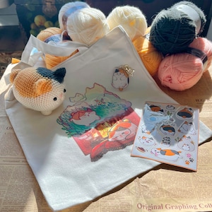 Puede incluir: Una bolsa de tela blanca con un diseño de gato de dibujos animados colorido. La bolsa está llena de ovillos de lana de varios colores, incluyendo rosa, blanco y negro. Hay un pequeño amuleto de gato unido a la bolsa.