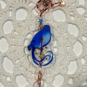 May include: A blue bird with a long tail sits on a purple staff with a crescent moon and stars. The bird is holding a small orb with a ring around it. The charm is attached to a gold chain.