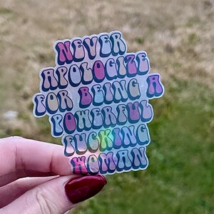 May include: A clear sticker with the text "NEVER APOLOGIZE FOR BEING A POWERFUL FUCKING WOMAN" in a retro font. The sticker has a holographic effect, with colors of pink, purple, and blue.
