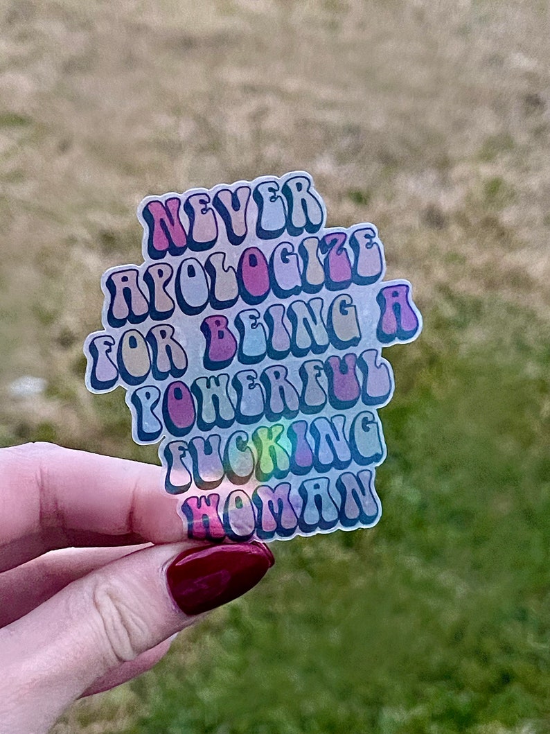 May include: A clear sticker with the text "NEVER APOLOGIZE FOR BEING A POWERFUL FUCKING WOMAN" in a retro font. The sticker has a holographic effect, with colors of pink, purple, and blue.