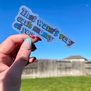 May include: A clear sticker with the text "That wasn't very cash money of you." in a rainbow of colors. The sticker is being held in a hand against a blue sky background.