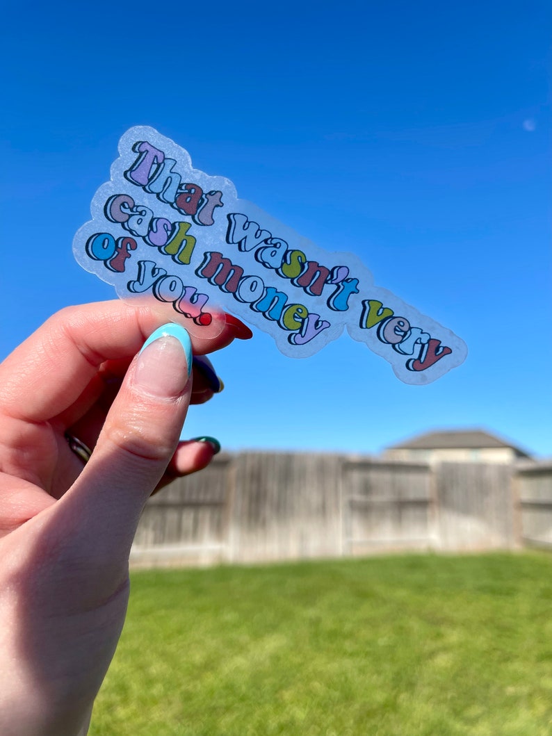 May include: A clear sticker with the text "That wasn't very cash money of you." in a rainbow of colors. The sticker is being held in a hand against a blue sky background.