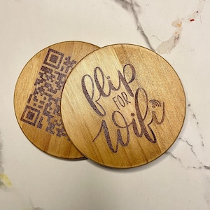 May include: Two round wooden coasters with a brown wood grain finish. One coaster has a black and white QR code and the other has the text "Flip for Wifi" in brown lettering.