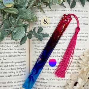 Pode incluir: Um marcador de livros de resina feito à mão com um gradiente de tons azuis e vermelhos, acentuado com uma borla rosa. O marcador de livros tem um acabamento brilhante e é projetado para amantes de livros. O fundo inclui um livro e vegetação.