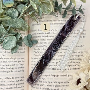 May include: A handmade bookmark with a swirling dark purple and white design. The bookmark has a white tassel and is resting on an open book. The bookmark is approximately 1 inch wide and 6 inches long.