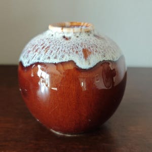 Op de afbeelding: Een kleine, ronde keramische vaas met een glanzende, bruine basis en een witte, getextureerde bovenkant. De vaas heeft een smalle opening en een gladde, afgeronde vorm. Het glazuur heeft een craquelé-effect, wat bijdraagt aan de vintage uitstraling.