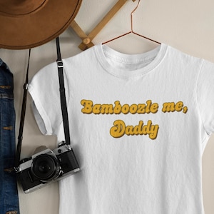May include: White t-shirt with the text "Bamboozle me, Daddy" in a retro yellow font.