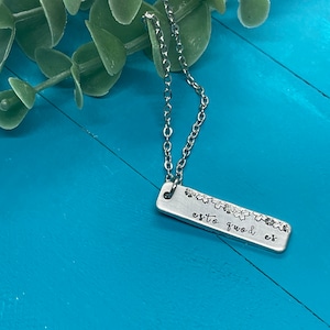 May include: A silver necklace with a rectangular pendant that reads "esto quod est" with a floral design. The necklace is on a blue wooden surface.
