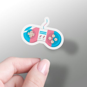 Puede incluir: Una pegatina de un mando de videojuegos retro con un esquema de color rosa, azul y blanco. El mando tiene una cruceta direccional, cuatro botones y las palabras "SELECT" y "START".