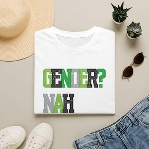 May include: A white short-sleeved t-shirt featuring the text GENDER? NAH printed in a blocky, distressed font. The letters alternate between shades of green, gray, and black, creating a textured, weathered appearance across the chest area.