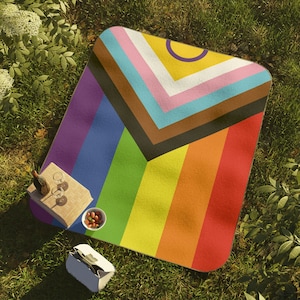 Könnte beinhalten: Eine rechteckige Picknickdecke mit Regenbogenfarben und dem Progress Pride Flaggen-Design. Auf dem Rasen befinden sich ein kleiner Holztisch, Weingläser und ein Picknickkorb. Das Design der Decke ist geometrisch.