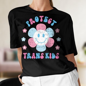 May include: Black t-shirt featuring a flower design with a smiling face and the words "PROTECT TRANS KIDS" in blue and pink. The flower petals are pink and light blue, with small flower accents.