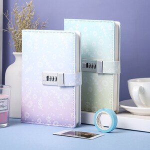 May include: Two pastel-coloured journals with combination locks. The journal on the left is purple and blue with a white floral pattern. The journal on the right is light blue and green with a white floral pattern. Both journals have a silver combination lock.