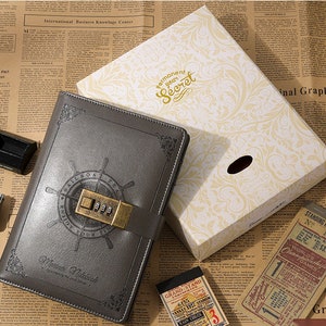 May include: A gray leather-bound journal with a combination lock and a compass design. The journal is closed and has the text "permanent your secret" on the box it is in.