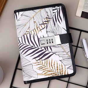 May include: A black leather-bound journal with a combination lock and a white and gold tropical leaf design.