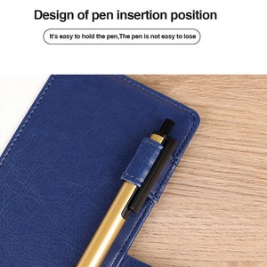 May include: A blue leather notebook with a gold pen inserted into a pen holder on the cover. The text "Design of pen insertion position" and "It's easy to hold the pen, The pen is not easy to lose" is visible on the image.