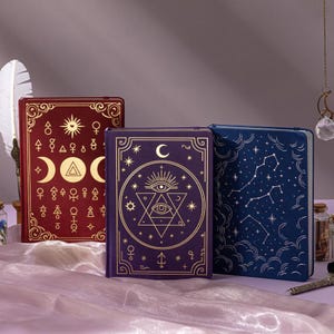 May include: Three hardback journals with celestial designs in burgundy, purple, and navy blue. The burgundy journal features a sun, moon, and triangle design. The purple journal has an eye and star design. The navy journal has a constellation design.