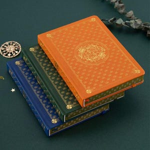 May include: Three hardback journals in blue, green, and orange, stacked. The covers have gold embossed floral designs and borders. The journals are closed, with a bookmark ribbon visible on the orange journal. The journals are approximately 15cm x 20cm.