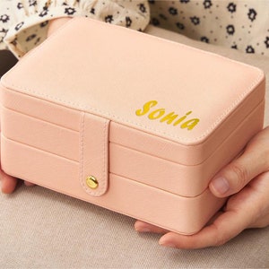 May include: A pink jewellery box with three layers and a gold clasp. The box has the name "Sonia" written on the top in gold lettering.