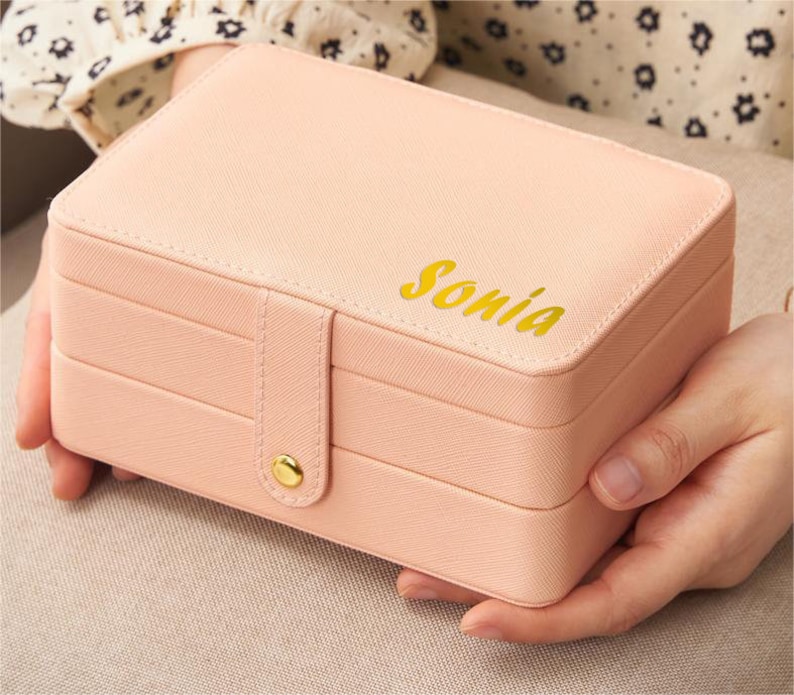 May include: A pink jewellery box with three layers and a gold clasp. The box has the name "Sonia" written on the top in gold lettering.