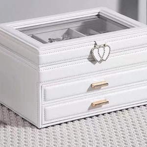 May include: White jewellery box with a glass top and two drawers. The box has a heart-shaped clasp and gold-coloured hardware.