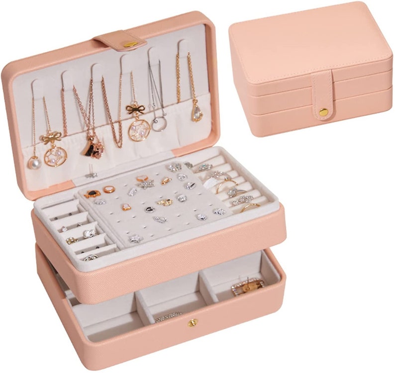 May include: A pink two-tiered jewellery box with a hinged lid and a pull-out drawer. The top tier has slots for necklaces, earrings, and rings. The bottom tier has compartments for rings and other small items.