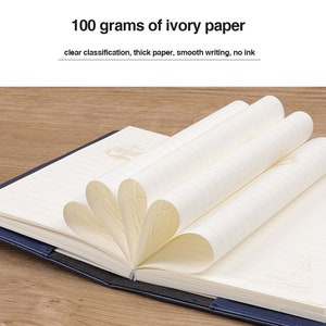 May include: A blue hardcover notebook with ivory paper. The notebook is open and the pages are fanned out to show the lined paper. The text on the cover reads "100 grams of ivory paper, clear classification, thick paper, smooth writing, no ink."