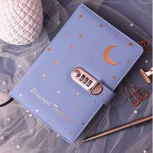 Puede incluir: Una agenda azul claro con un diseño de luna y estrellas doradas. La agenda tiene una cerradura de combinación y las palabras "Beautiful Tomorrow" escritas en la portada.
