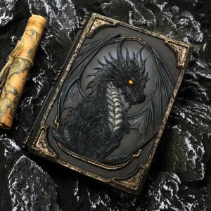 以下が含まれることがあります： 黒革製のジャーナルで、精巧なエンボス加工が施されたドラゴンデザインが施されています。ドラゴンは金色のアクセントと輝く黄色の目をしています。