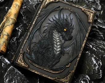 May include: A black leather-bound journal with a detailed embossed dragon design. The dragon has gold accents and glowing yellow eyes.