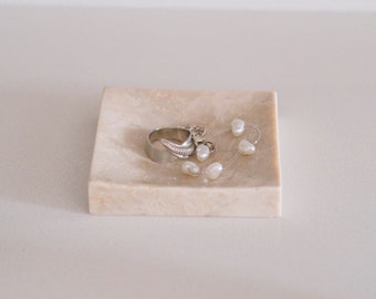 May include: A small, square, light beige marble dish with a silver ring and white pearl-like beads inside.