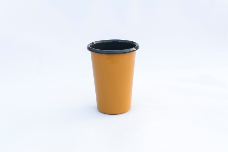 Yellow enamel tumbler cup with anthracite rim, placed on a white surface, with a plain background.