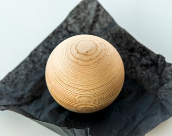 May include: A light brown wooden sphere with a smooth, textured surface. The sphere is sitting on a black piece of fabric.
